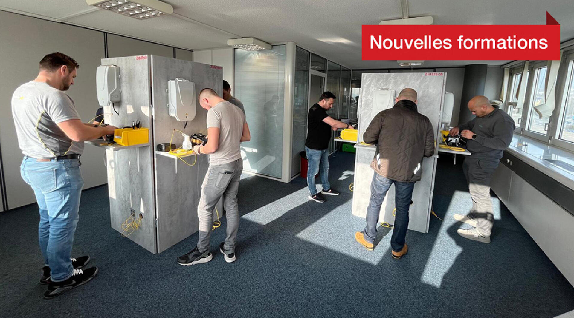 Cinq hommes s'exercent au câblage sur des panneaux de formation dans une salle de classe lumineuse. La scène comporte de grandes fenêtres, une moquette bleue et une bannière rouge en français indiquant "Nouvelles formations" dans le coin supérieur droit.