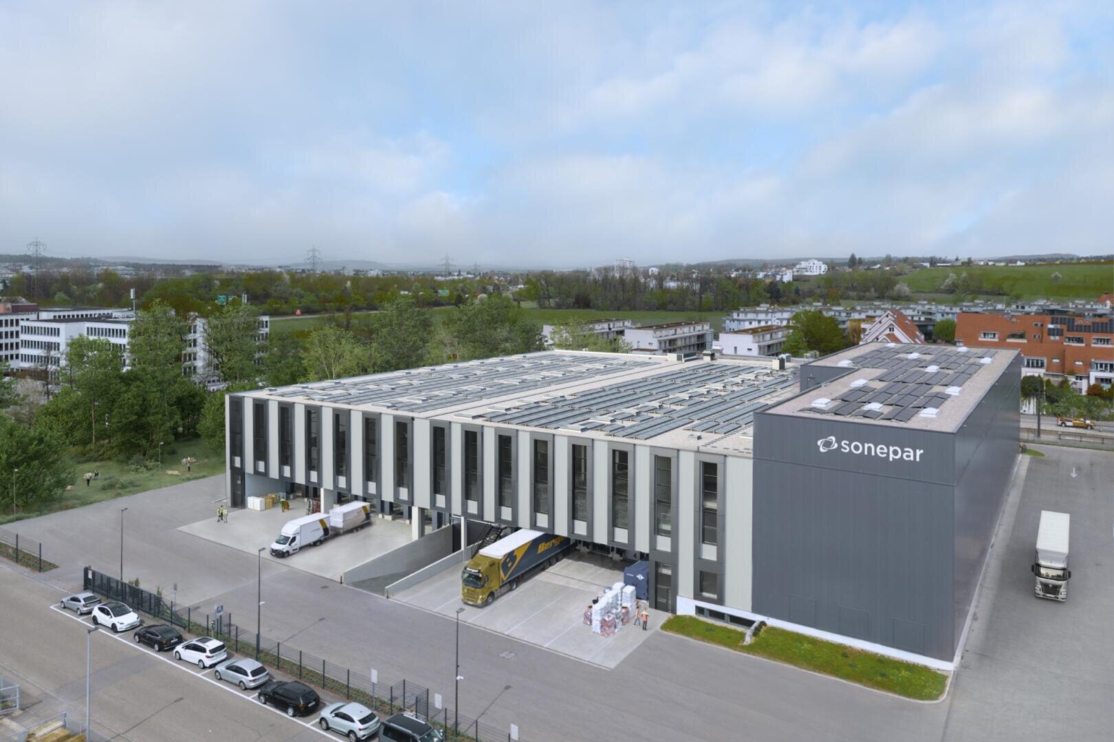 Ein modernes, mehrstöckiges Gebäude von Sonepar mit Solarmodulen auf dem Dach, umgeben von Grünflächen und Straßen. Mehrere Lkw und ein Bus stehen in der Nähe der Laderampen, während im Vordergrund Autos entlang einer Straße geparkt sind.
