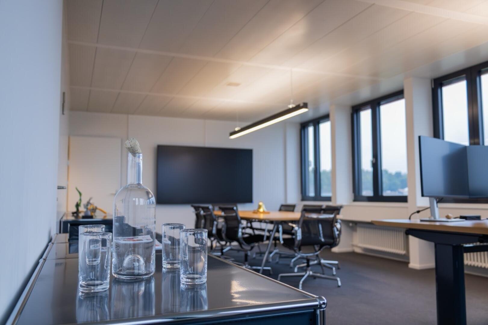 Un bureau moderne doté de grandes fenêtres, d'une table de réunion avec des chaises, de deux écrans plats et d'un buffet sur lequel sont posés une bouteille en verre et plusieurs verres vides. L'espace est propre et bien éclairé.
