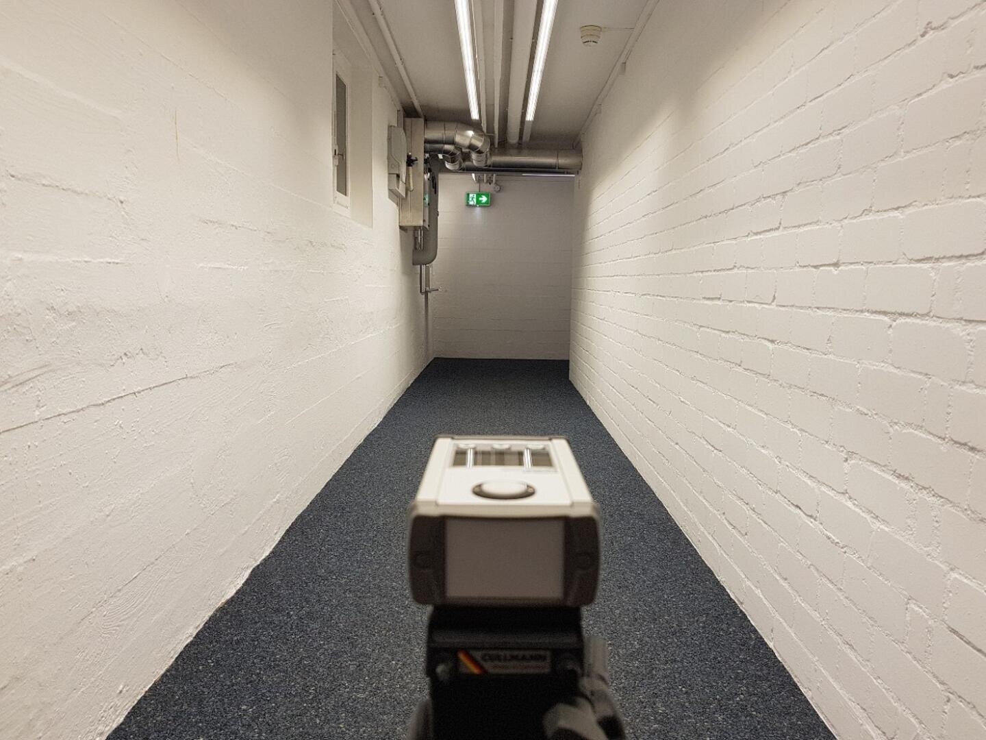 Un appareil se trouve à l'entrée d'un long couloir étroit aux murs de briques blanches, avec un tapis bleu et des tuyaux au sol. Un panneau d'issue de secours vert éclairé est visible à l'autre extrémité.