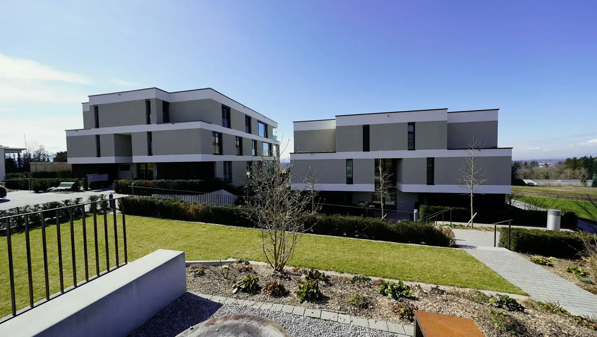 Deux immeubles d'habitation modernes, rectangulaires, aux toits plats, se trouvent côte à côte, entourés de pelouses vertes, d'arbustes et de trottoirs pavés, sous un ciel bleu limpide.