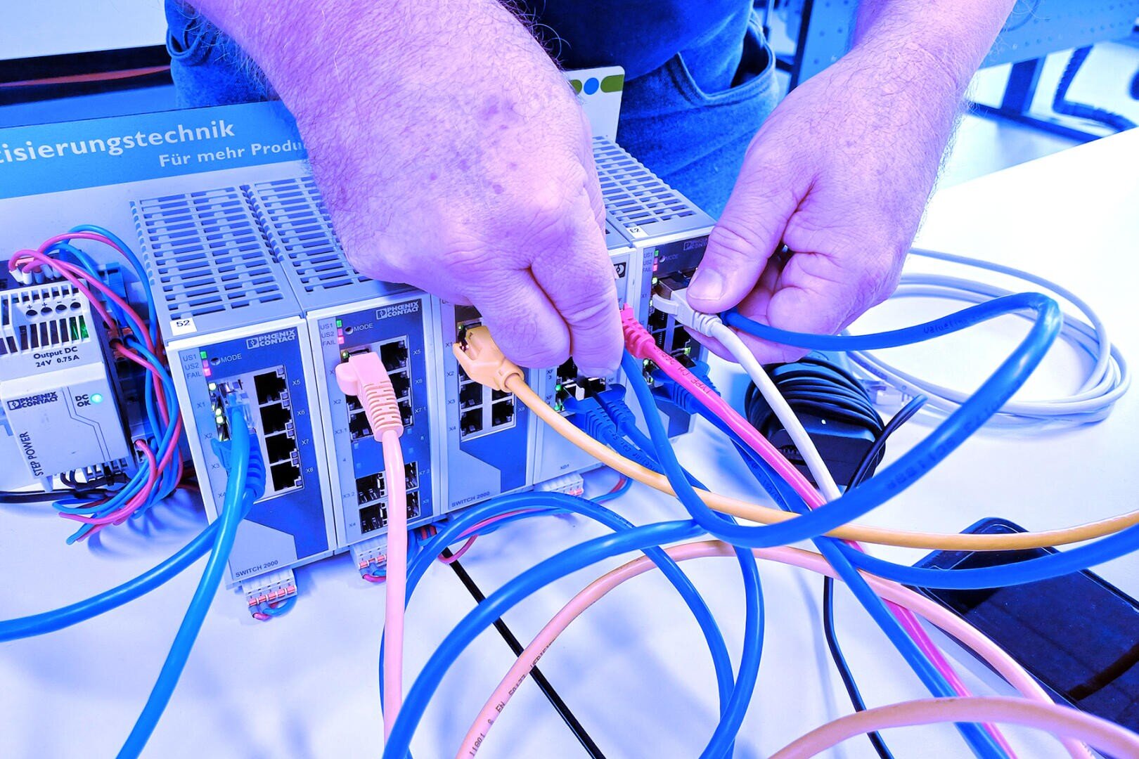 Hände, die mehrere bunte Ethernet-Kabel an Netzwerk-Switches auf einem Schreibtisch anschließen, wobei verschiedene Netzwerkgeräte zu sehen sind. Die Szene deutet auf eine technische Installation, Wartung oder Netzwerkkonfiguration hin.