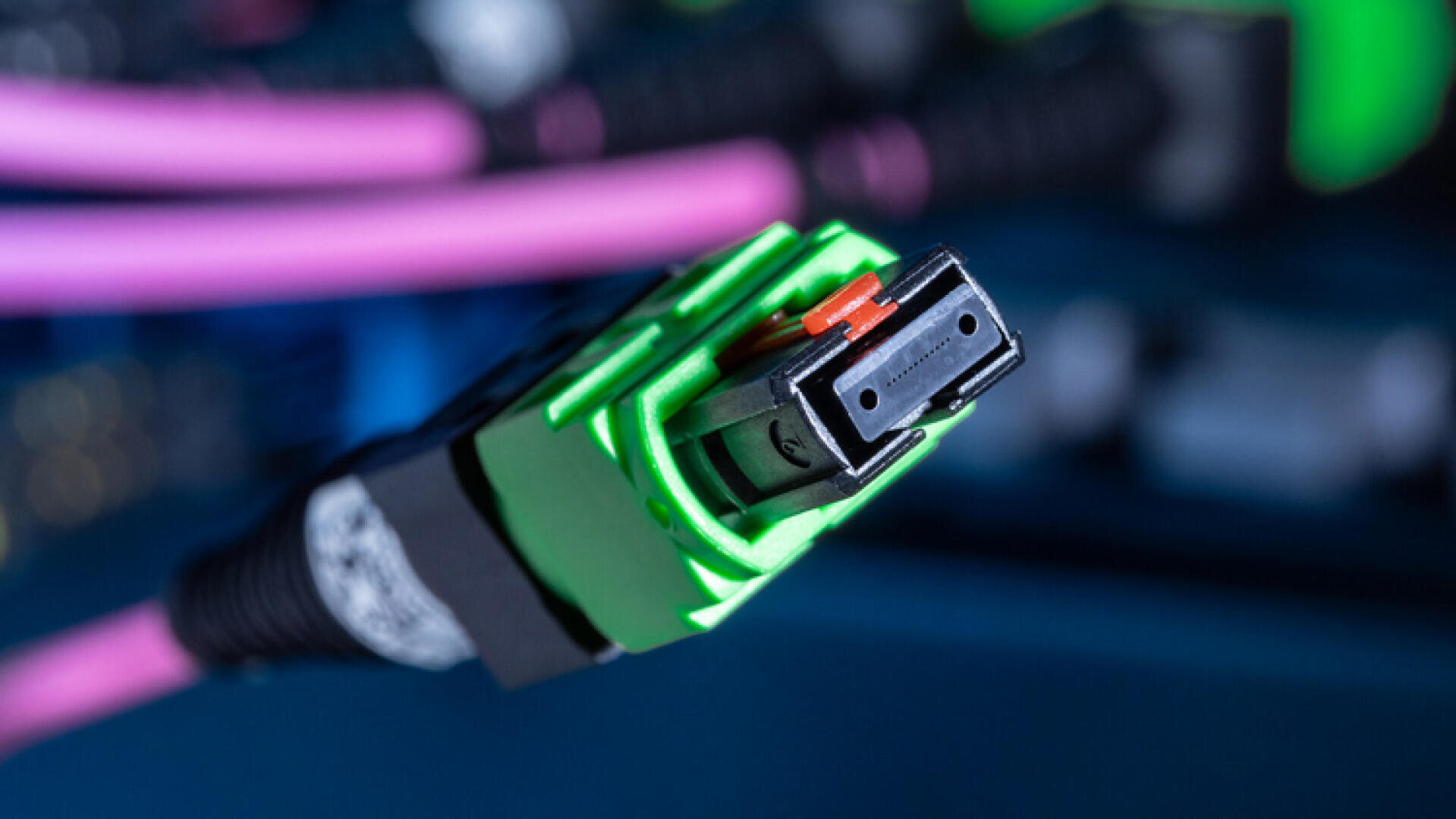Gros plan sur un connecteur de fibre optique vert auquel sont raccordés des câbles violets, montrant l'extrémité rectangulaire du connecteur et les détails fins des composants internes. L'arrière-plan est flou.