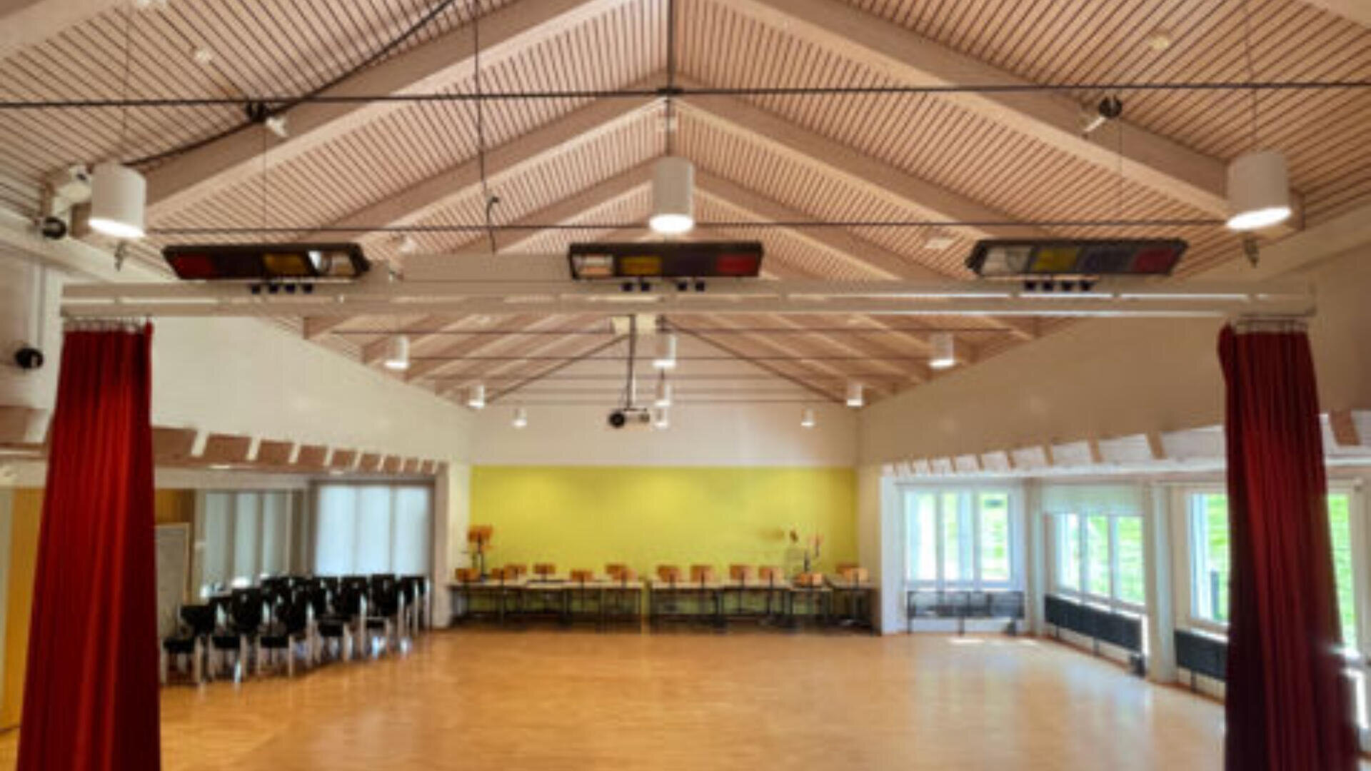 Ein geräumiger, heller Raum mit Holzboden, roten Vorhängen, hohen Deckenbalken, schwarzen Stühlen auf der linken Seite und großen Fenstern an der rechten Wand, durch die Tageslicht fällt.