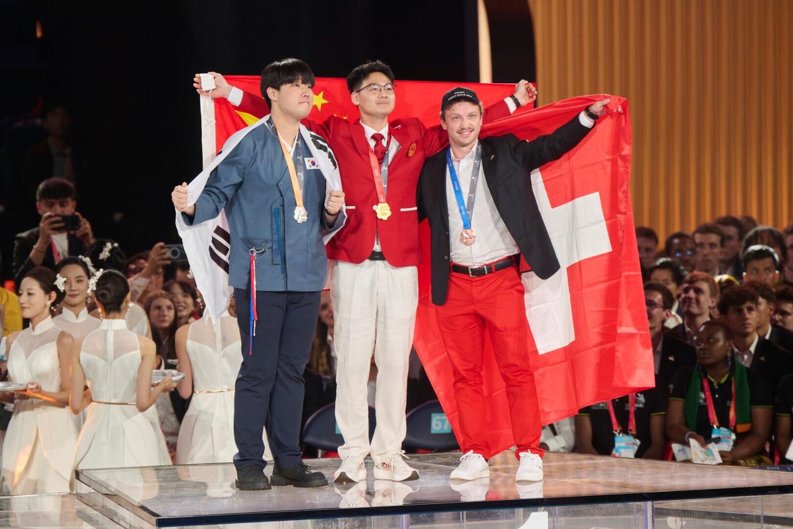 Drei Medaillengewinner stehen auf einem Podium und halten jeweils die Flagge ihres Landes in der Hand: Südkorea (links, Silber), China (Mitte, Gold) und die Schweiz (rechts, Bronze). Hinter ihnen schaut das Publikum zu.