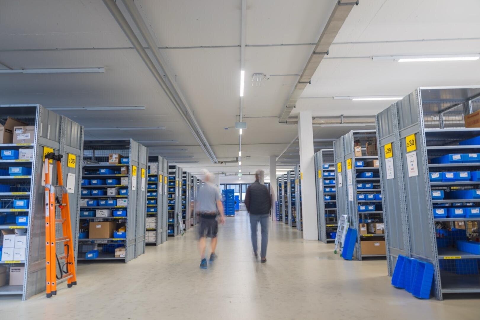 Un entrepôt très bien éclairé, avec des rayonnages métalliques remplis de bacs et de cartons bleus. Deux personnes avancent dans l'allée centrale, sous un éclairage industriel et au milieu de panneaux indiquant les différentes zones de stockage.