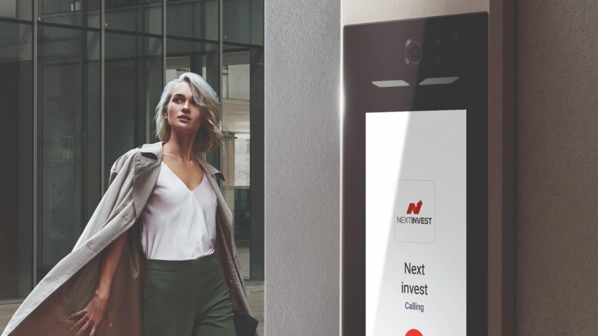Eine Frau mit kurzen blonden Haaren und einem hellen Mantel geht an einem modernen Gebäude vorbei; neben ihr befindet sich eine Sprechanlage mit dem NextInvest-Logo und einer Ruftaste.
