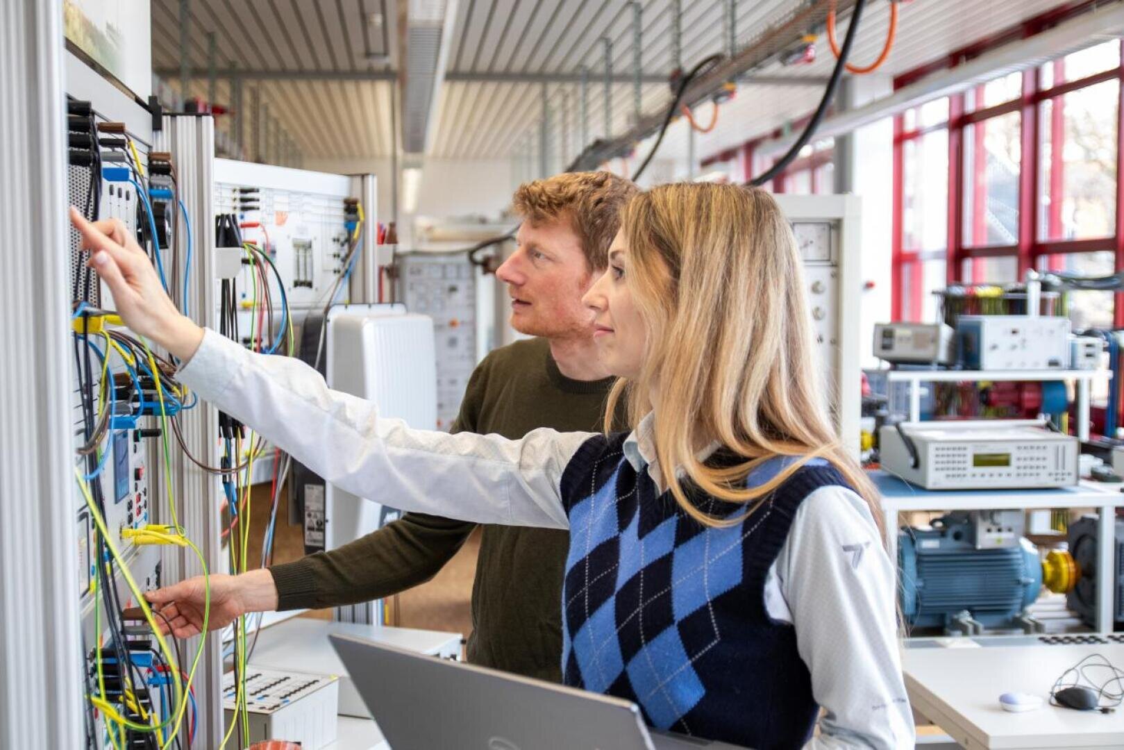 Zwei Personen stehen in einem hellen Labor und arbeiten an einer Elektronikplatine mit verschiedenen Kabeln. Eine von ihnen zeigt auf das Gerät, während die andere, die einen Laptop in der Hand hält, aufmerksam zusieht. Im Hintergrund sind technische Geräte und Maschinen zu sehen.