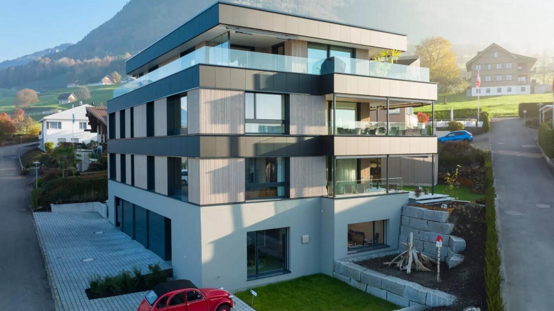 Un immeuble d'appartements moderne de trois étages avec des balcons en verre et des accents en bois, situé sur un terrain d'angle dans une zone suburbaine avec des collines verdoyantes et quelques maisons voisines. Une voiture rouge est garée devant.