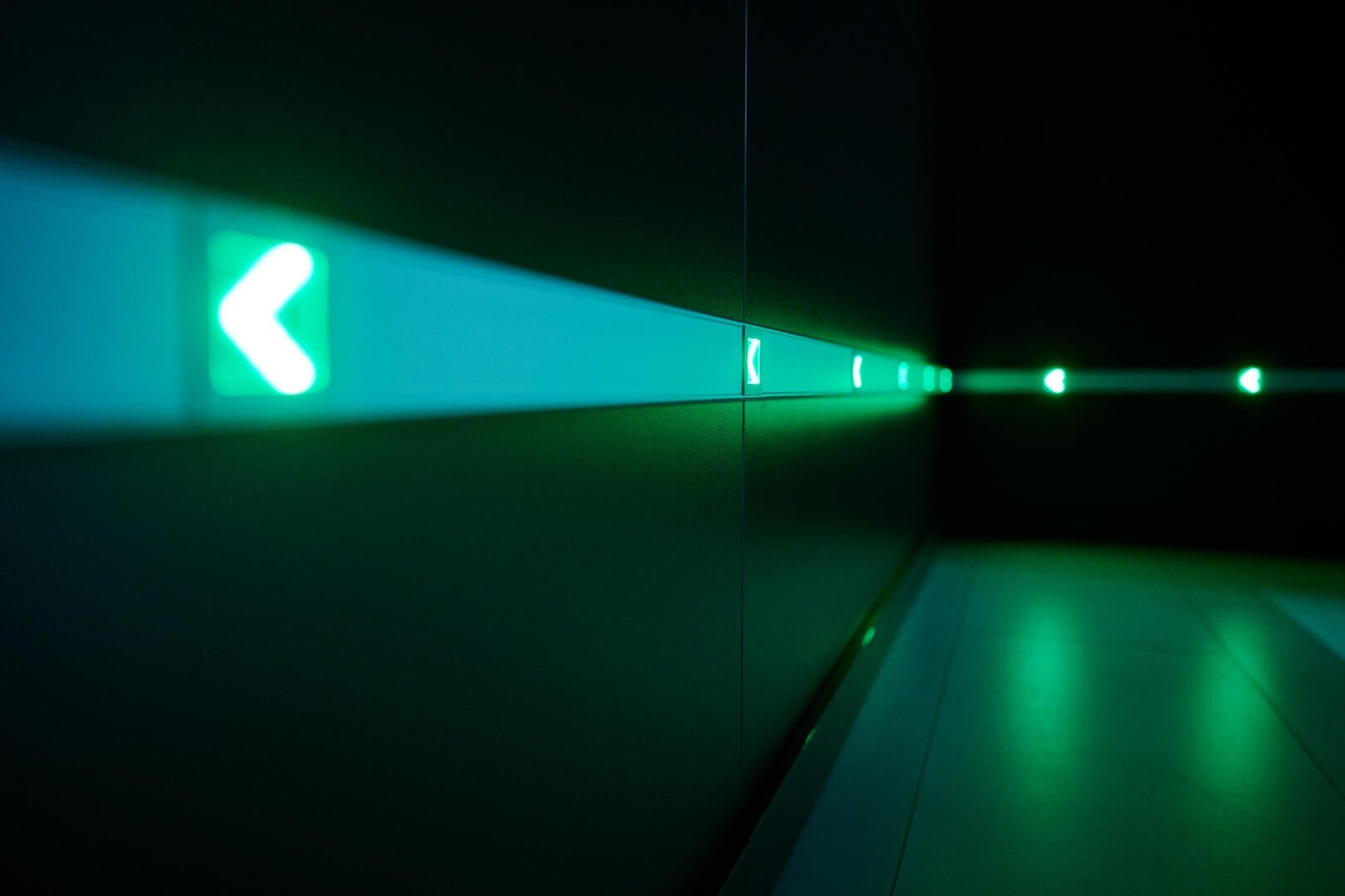 Ein dunkler Flur mit einer Reihe leuchtender grüner Pfeilschilder an der Wand, die alle nach links zeigen und eine futuristische und minimalistische Atmosphäre schaffen.