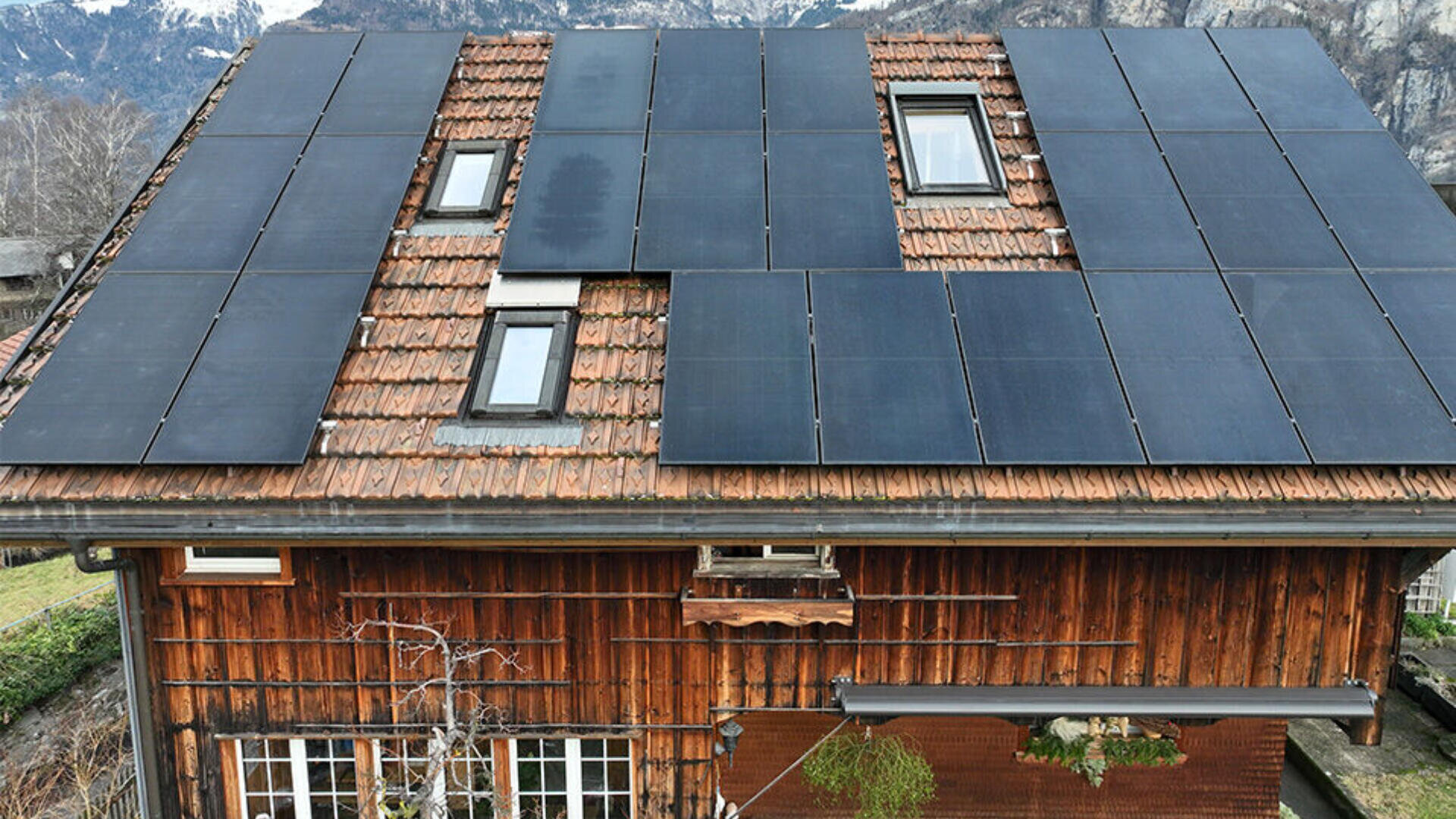 In einem Holzhaus mit rotem Ziegeldach sind mehrere schwarze Sonnenkollektoren installiert, die mehrere Oberlichter umgeben. Im Hintergrund sind verschneite Berge und blattlose Bäume zu sehen.