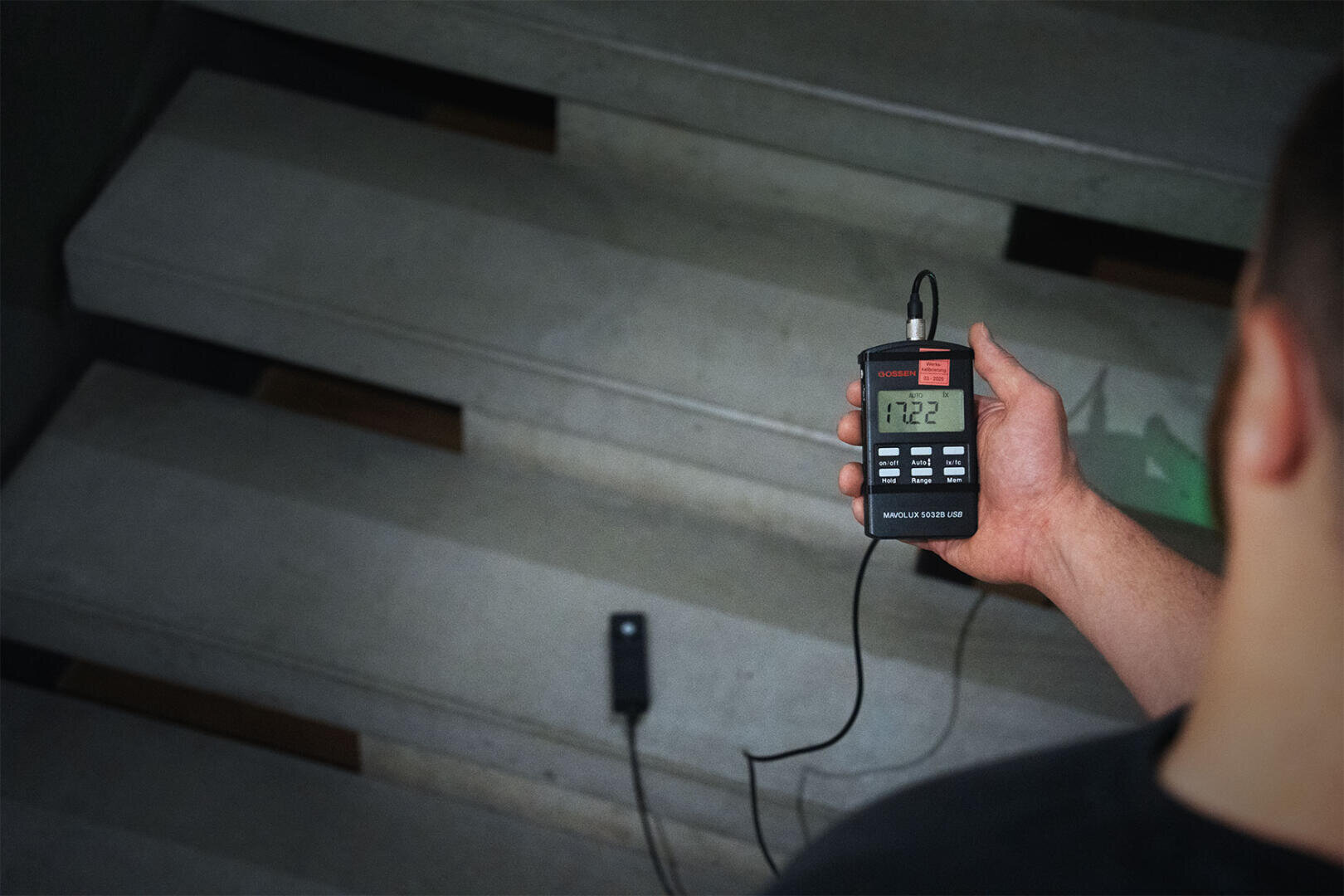 Eine Person hält ein digitales Belichtungsmessgerät in der Hand, das einen Wert von 112,2 anzeigt, während sie die Helligkeit auf einer Treppe misst, wobei der Sensor auf einer Treppenstufe angebracht ist.