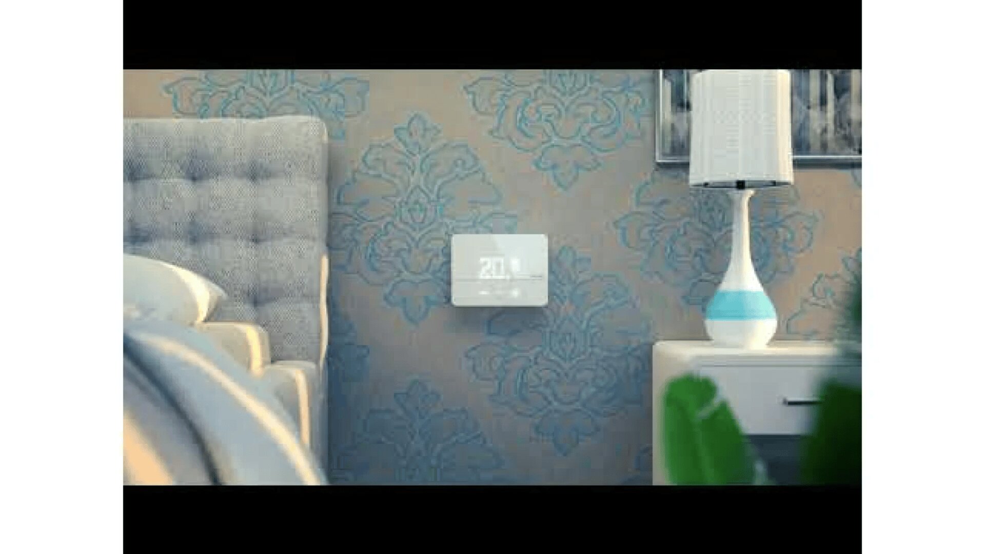 Ein digitaler Thermostat ist an einer blau gemusterten Wand neben einem grauen Polsterbett und einem weißen Nachttisch mit einer weiß-blauen Lampe in einem schummrig beleuchteten Schlafzimmer angebracht.