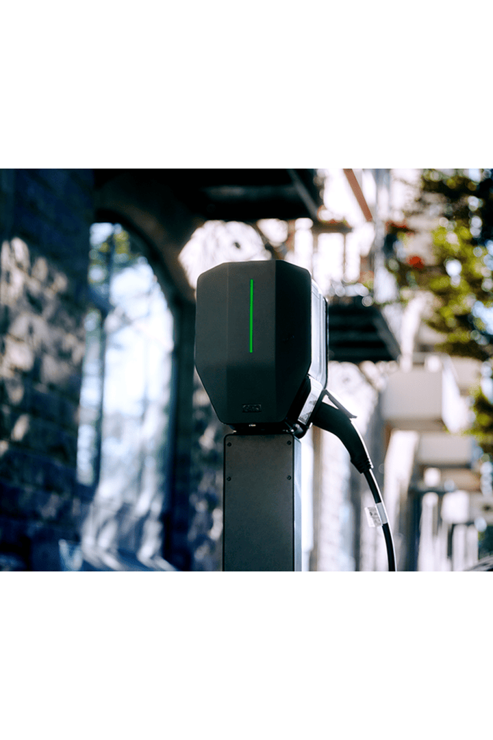 Eine Nahaufnahme einer Ladestation für Elektrofahrzeuge im Freien mit einer grünen Leuchtanzeige und einem angeschlossenen Ladekabel vor einem unscharfen städtischen Hintergrund.