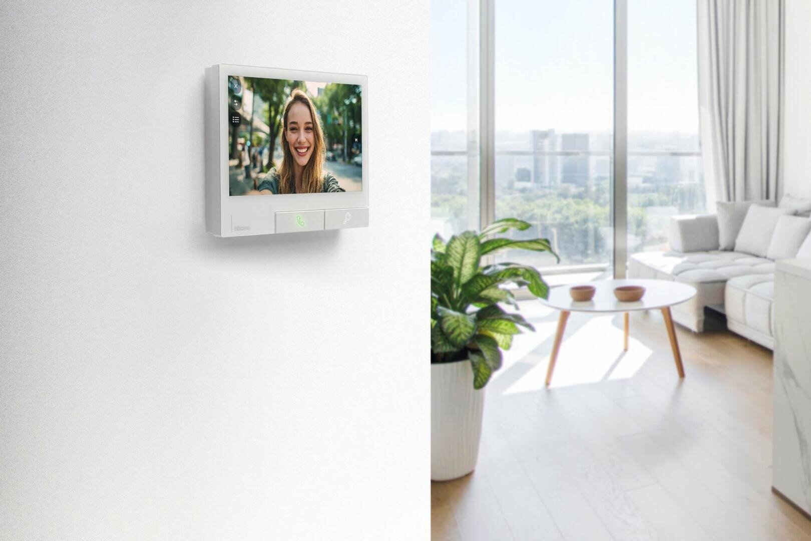 Une femme souriante apparaît sur l'écran de l'interphone vidéo installé sur un mur blanc. Le salon moderne et lumineux au fond a de grandes fenêtres, un canapé blanc, une plante, une table basse ronde et une vue sur la ville à l'extérieur.