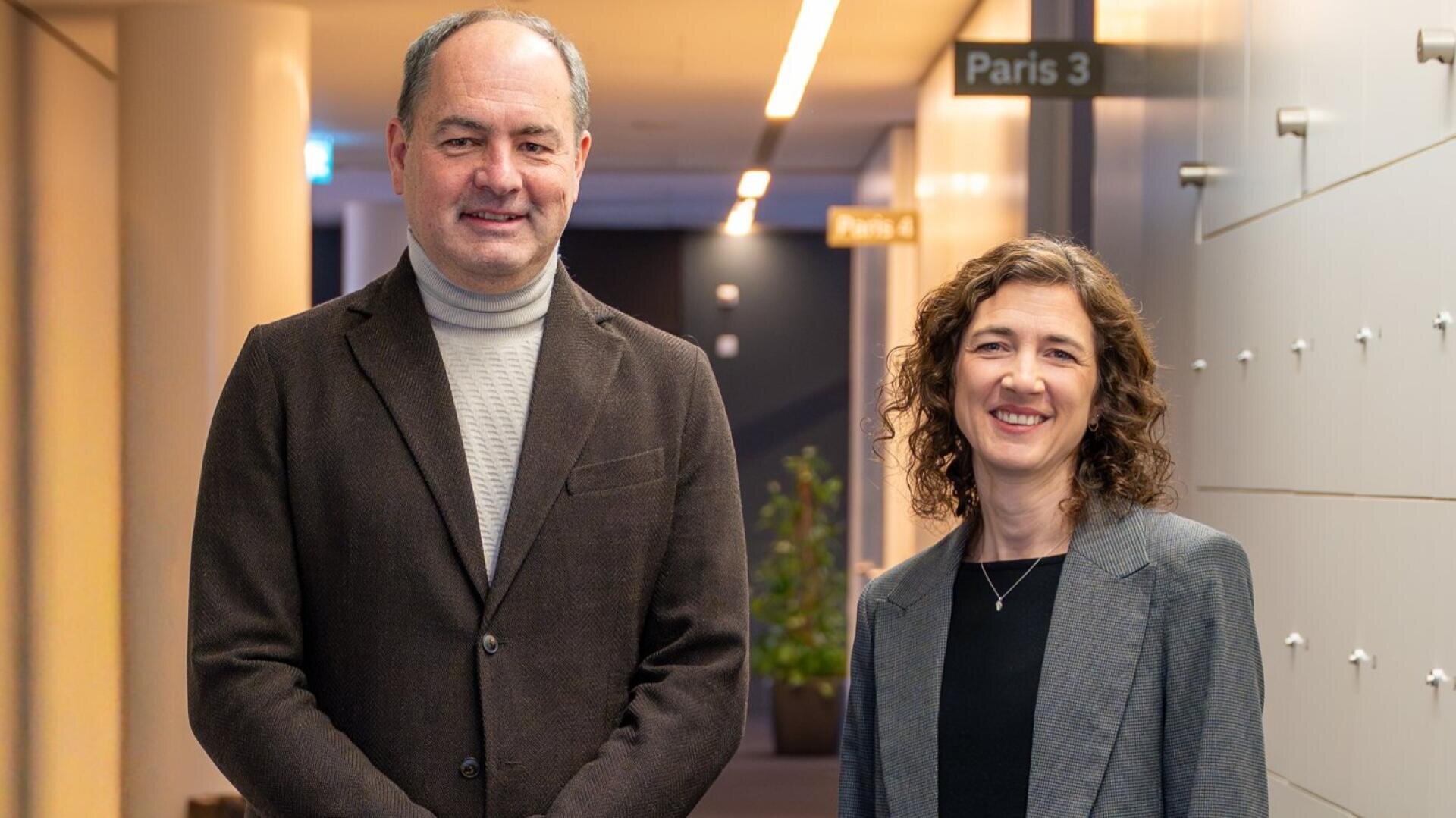 Deux personnes en tenue de travail se tiennent dans un couloir moderne avec des panneaux marqués Paris 3 et Paris 4 visibles à l'arrière-plan. Les deux personnes sourient et regardent la caméra.