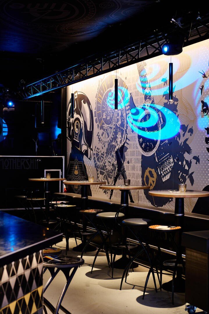 Eine schummrig beleuchtete Bar mit hohen Tischen und Hockern besticht durch ein großes Wandgemälde mit Robotern und abstrakten Motiven, das von blauen Neonlichtern beleuchtet wird und eine moderne, futuristische Atmosphäre schafft.
