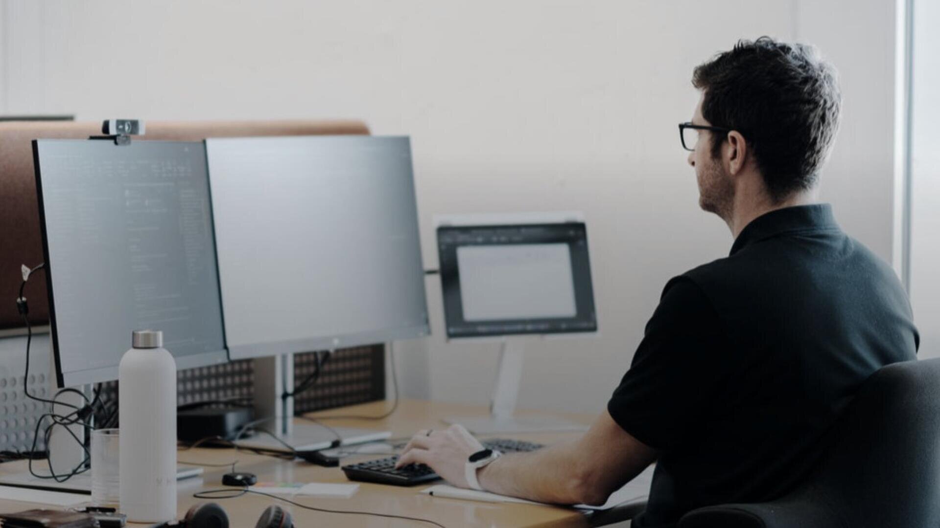 Eine Person mit Brille und schwarzem Hemd arbeitet an einem Schreibtisch mit zwei Bildschirmen und einem Tablet. Auf dem Schreibtisch stehen außerdem eine weiße Wasserflasche und Kopfhörer. Die Person scheint sich ganz auf die Bildschirme zu konzentrieren.