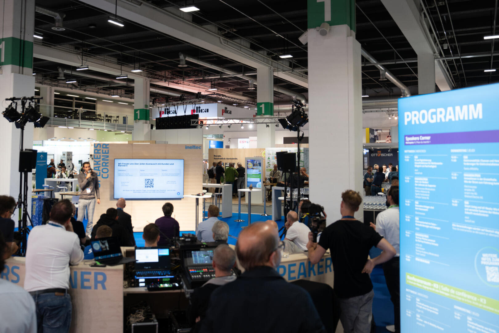 Eine Gruppe von Personen nimmt an einer Präsentation auf einer Technologiemesse teil. Es gibt eine Bühne mit einem Bildschirm, auf dem eine Folie gezeigt wird, eine große blaue Zeitplantafel im Vordergrund und verschiedene Stände und Geräte im Hintergrund.