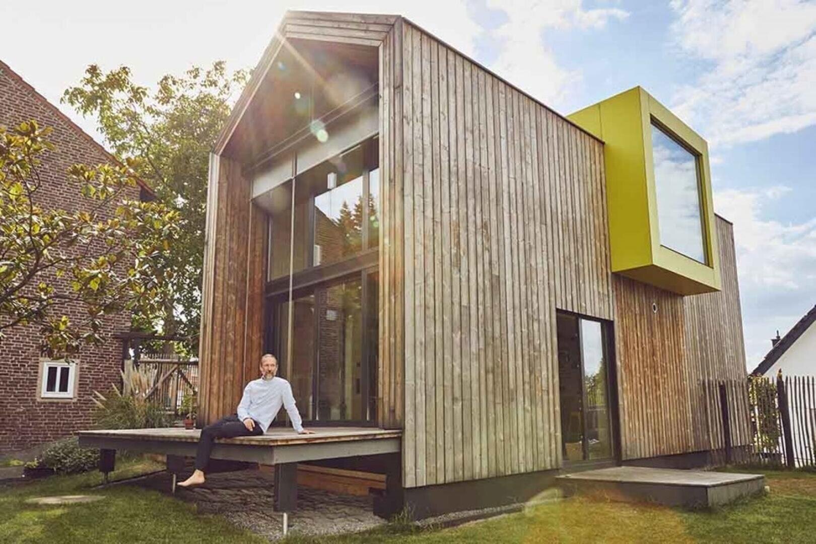 Ein Mann sitzt auf der hölzernen Veranda eines modernen, kleinen Hauses mit Holzverkleidung und großen Fenstern. Das Haus hat ein charakteristisches gelb umrahmtes Fenster und ist von Bäumen und benachbarten Häusern umgeben.