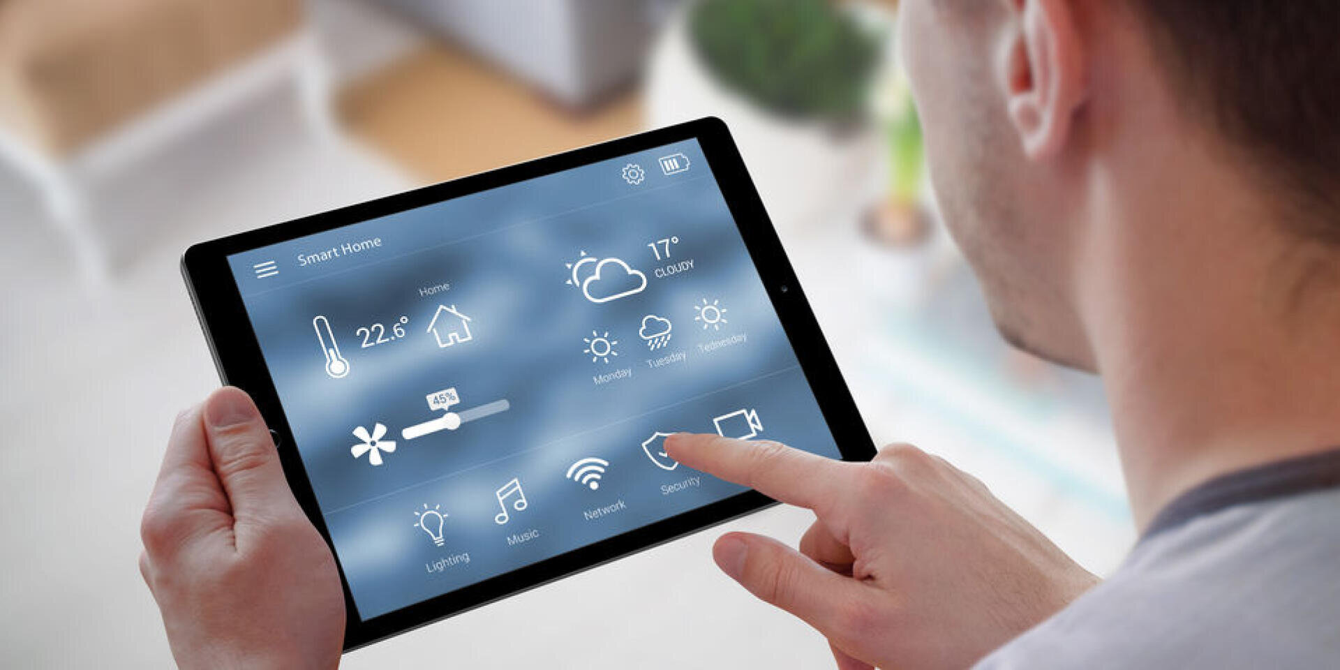 Eine Person hält und bedient ein Tablet, auf dem eine Schnittstelle für die Steuerung des intelligenten Zuhauses angezeigt wird, mit Symbolen für Temperatur, Wetter, Beleuchtung, Musik, Netzwerk, Sicherheit und andere Hausautomatisierungsfunktionen.