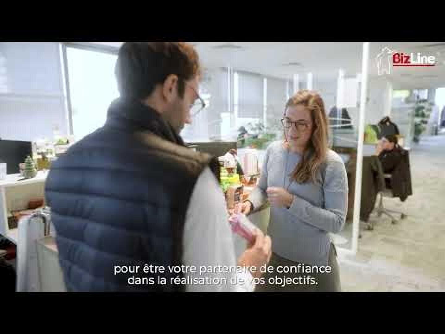 Un homme et une femme se tiennent dans un bureau moderne et lumineux, souriant et discutant. L'homme tient des papiers et la femme un stylo. Un texte en français et le logo BizLine apparaissent sur l'image. D'autres personnes travaillent à des bureaux à l'arrière-plan.