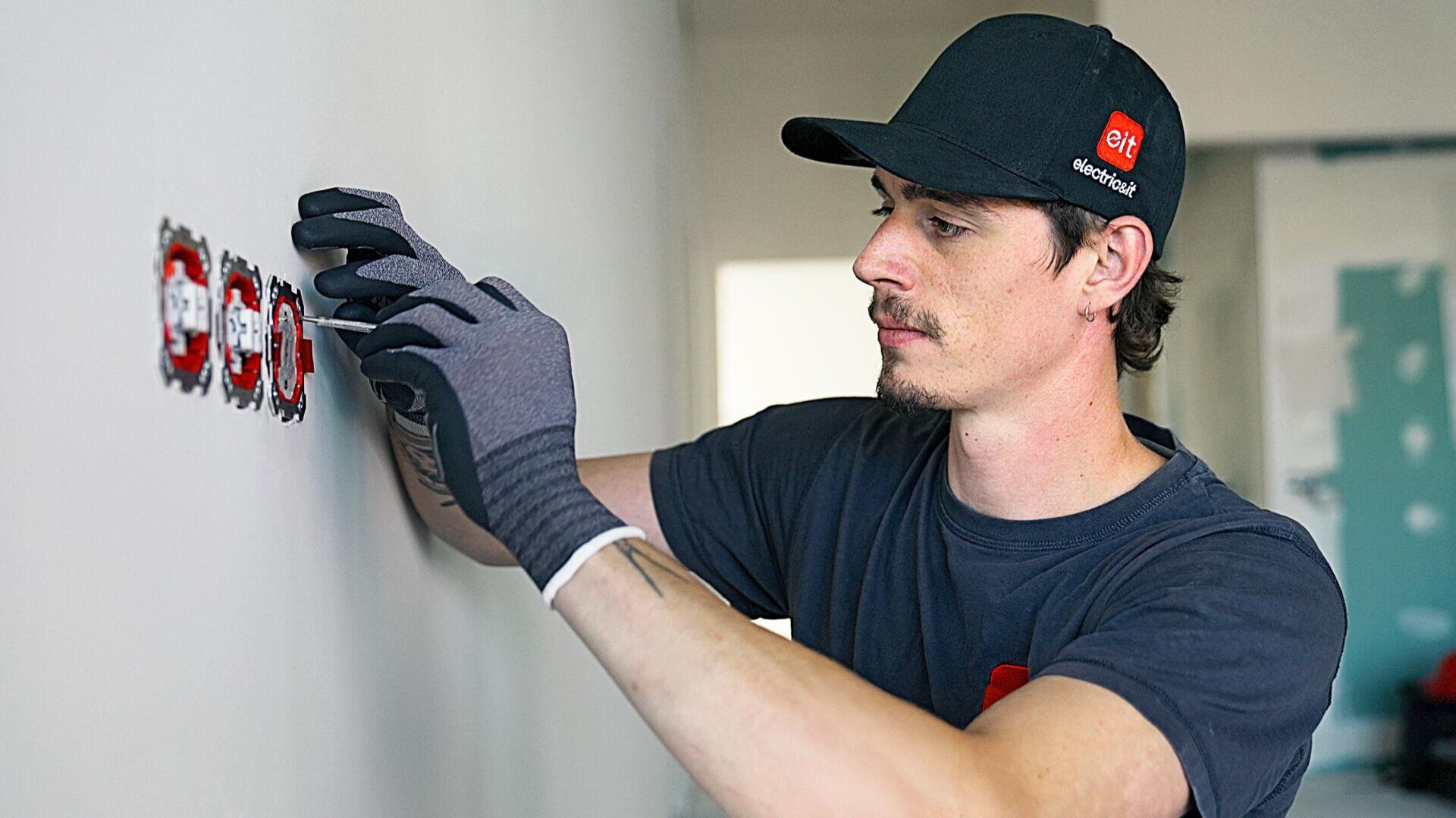 Un homme portant des gants et une casquette noire installe des prises électriques dans un mur. Il s'applique à bien aligner les prises, tandis qu'à l'arrière-plan on aperçoit une pièce en cours d'aménagement.
