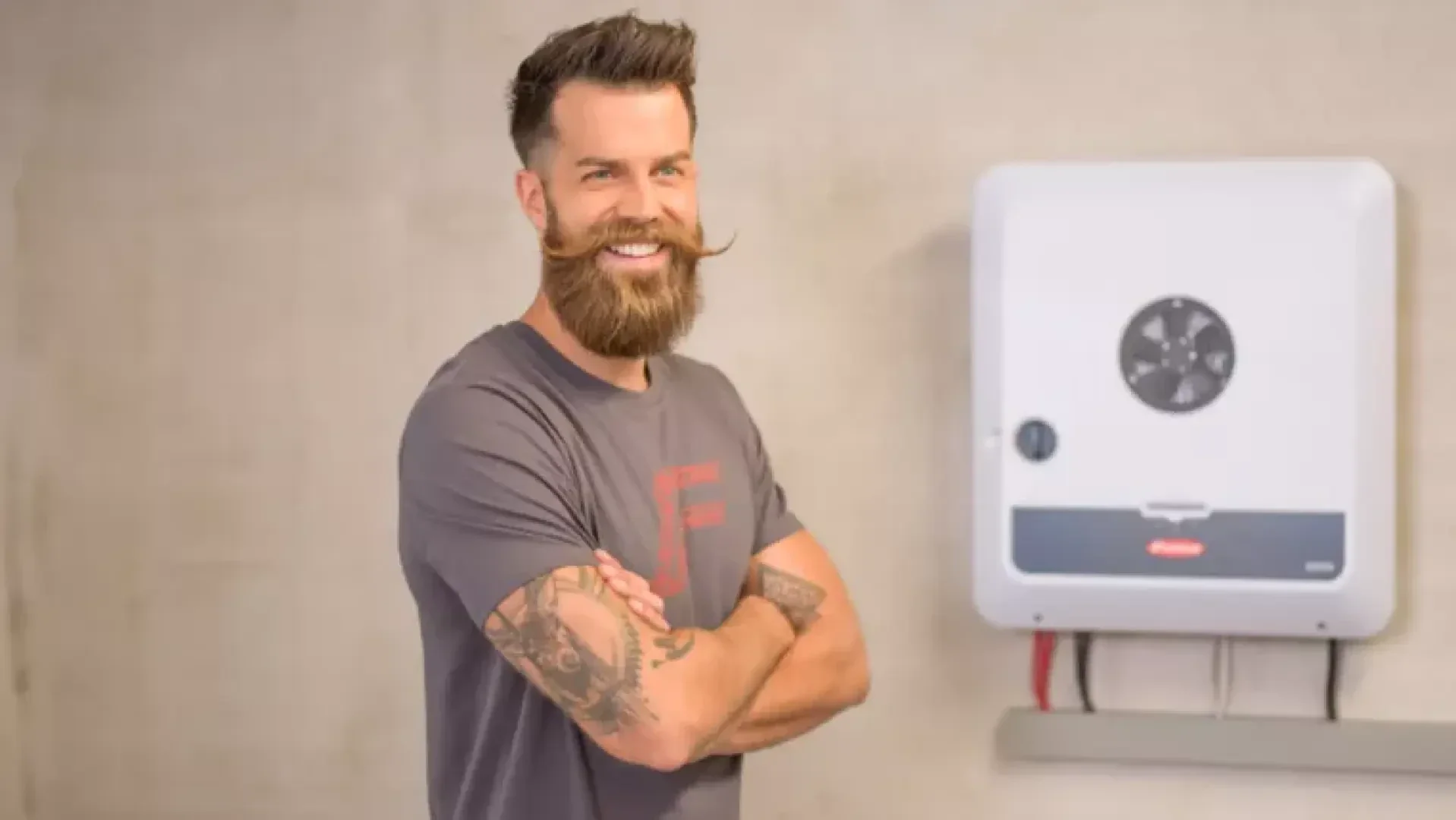 Un homme barbu, portant un t-shirt gris, se tient debout, les bras croisés, et sourit devant un appareil électrique blanc fixé au mur.