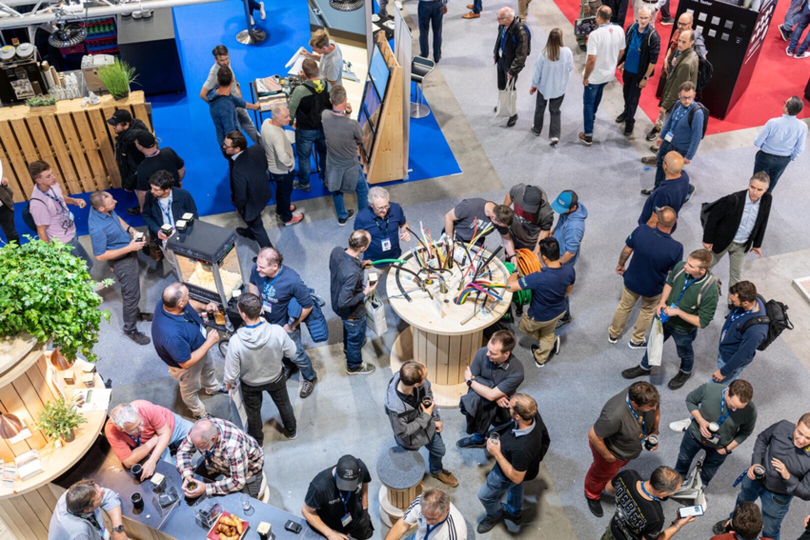 Bei einer überfüllten Veranstaltung in einer Halle versammeln sich Menschen um Ausstellungstische mit Kabeln und elektronischen Geräten, tauschen sich aus, unterhalten sich und betrachten die Exponate. Die Atmosphäre ist geschäftig und informell, mit Ständen und Topfpflanzen.