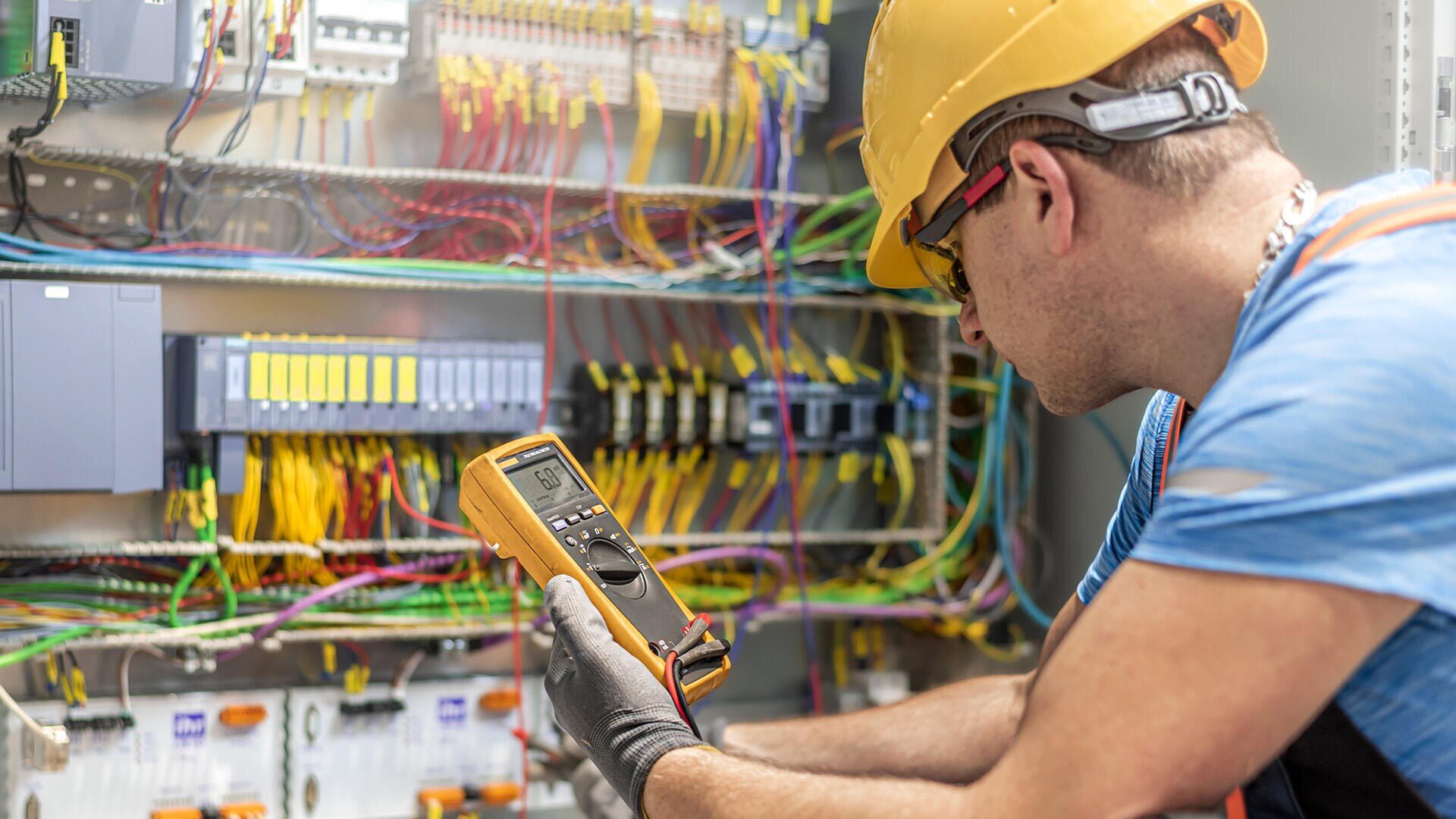 Ein Elektriker, der einen Schutzhelm und Handschuhe trägt, überprüft mit einem Digitalmultimeter die Verkabelung in einem offenen Schaltschrank, der mit bunten Kabeln und Bauteilen gefüllt ist.