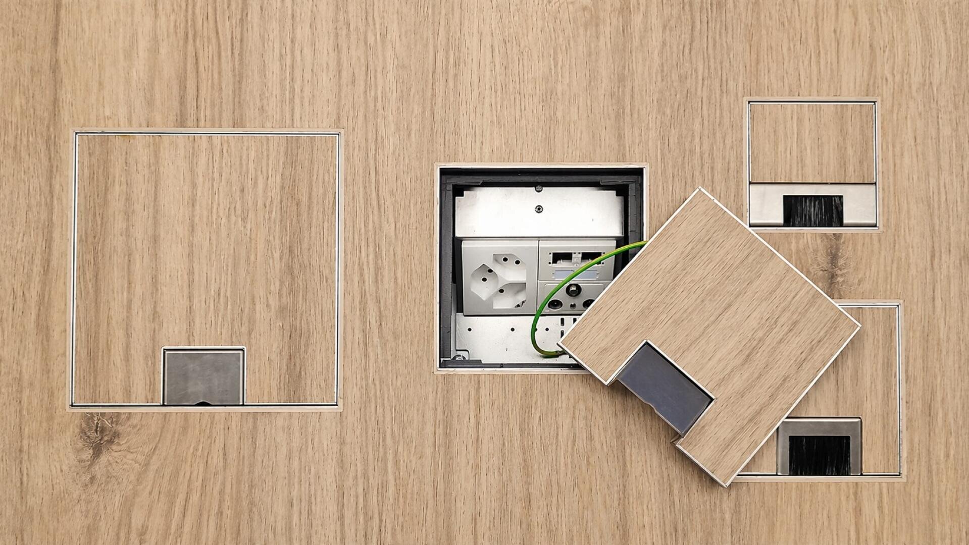 Un panneau en bois avec plusieurs découpes rectangulaires, dont l'une montre des prises et des câbles derrière un cache en bois partiellement ouvert. Les autres découpes sont fermées par des caches en bois assortis.
