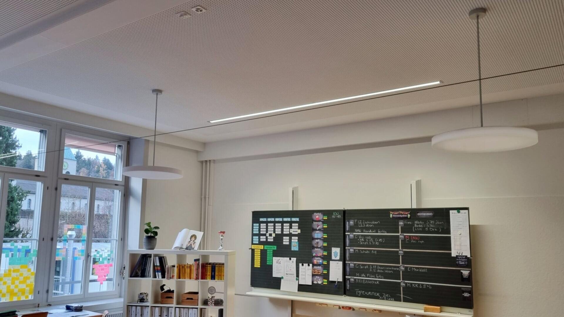 Une salle de classe dotée de grandes fenêtres, d'un tableau noir couvert de notes et d'emplois du temps, de murs blancs, d'étagères et de suspensions. La lumière naturelle pénètre par le côté gauche.