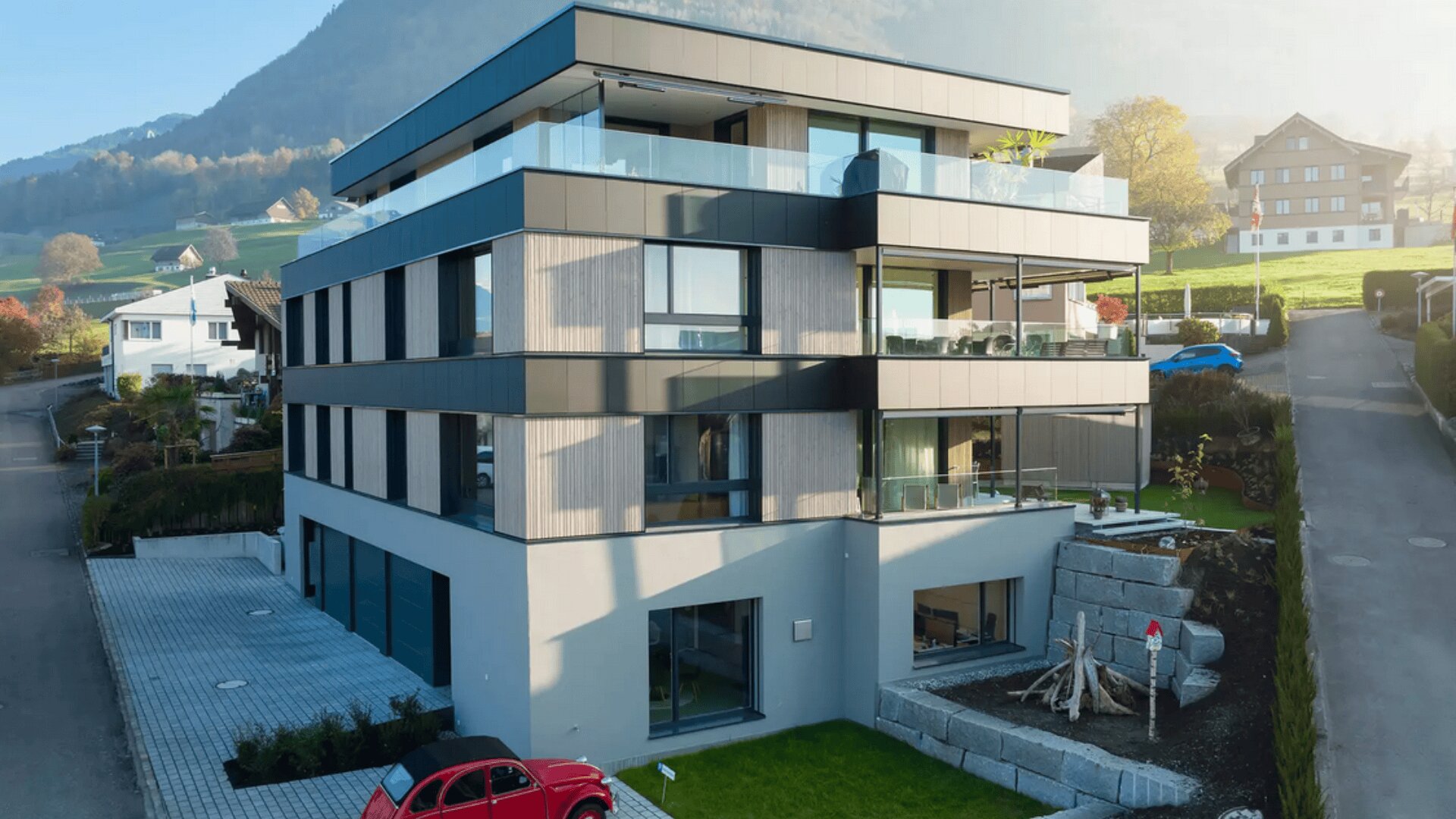 Ein modernes dreistöckiges Wohngebäude mit Glasbalkonen, das an einem grasbewachsenen Hang mit Bergen im Hintergrund liegt. Ein rotes Auto ist davor geparkt, und die Szene wird von sanftem Tageslicht beleuchtet.