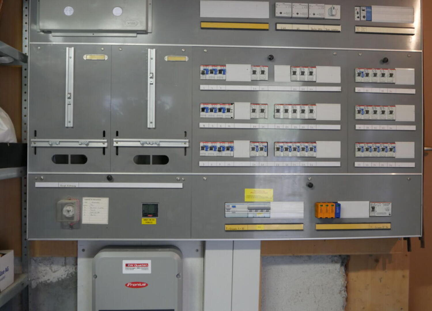 Eine große elektrische Schalttafel mit mehreren Schaltern, Leistungsschaltern, Aufklebern und Messgeräten, die an einer Wand montiert ist, mit einem grauen Wechselrichtergerät unten links.