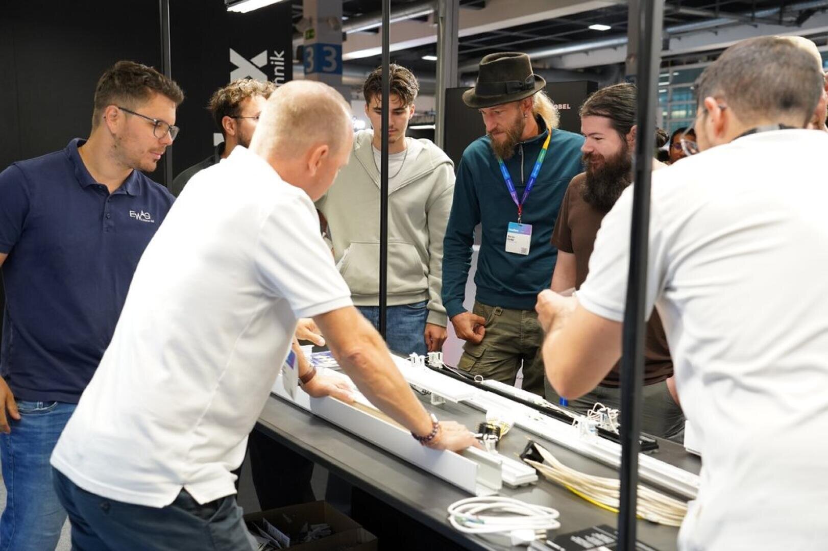 Eine Gruppe von Männern steht um einen Tisch herum und verfolgt bei einer Indoor-Veranstaltung oder Messe eine Vorführung mit elektrischen Geräten und Leuchten.