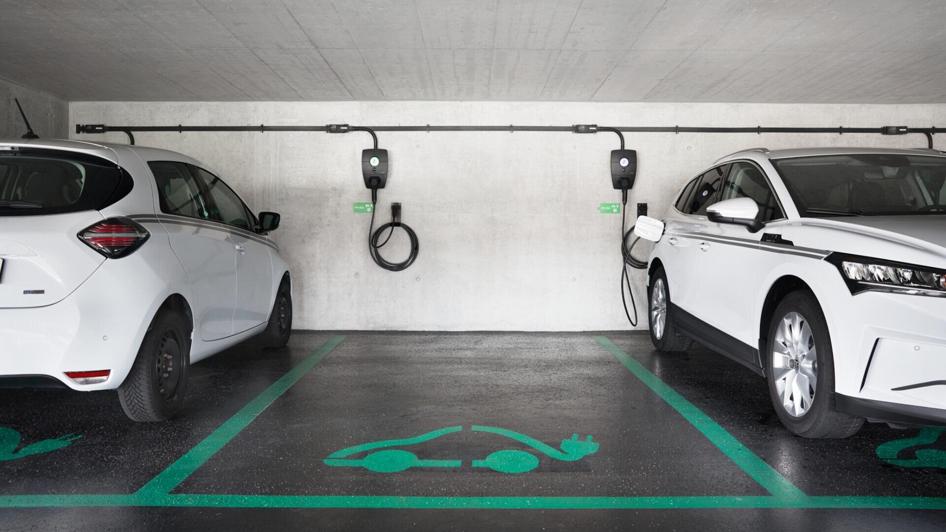 Zwei weiße Elektroautos stehen an Ladestationen in einer Garage und sind jeweils an Wandladegeräte angeschlossen. Grüne Markierungen auf dem Boden kennzeichnen die ausgewiesenen Stellplätze für Elektrofahrzeuge.