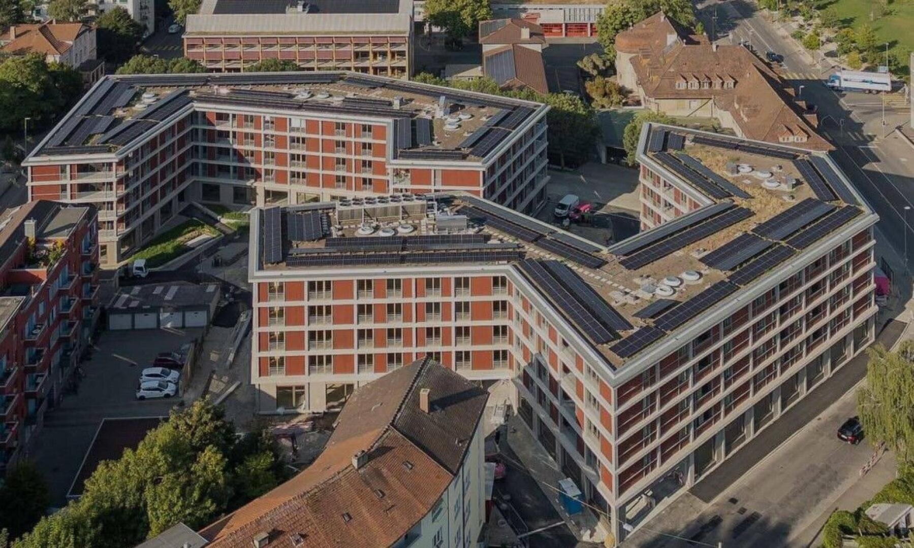 Luftaufnahme eines großen, modernen Wohngebäudes mit Sonnenkollektoren auf dem Dach, die in einem Zickzackmuster angeordnet und von Straßen, Bäumen und anderen Gebäuden umgeben sind.