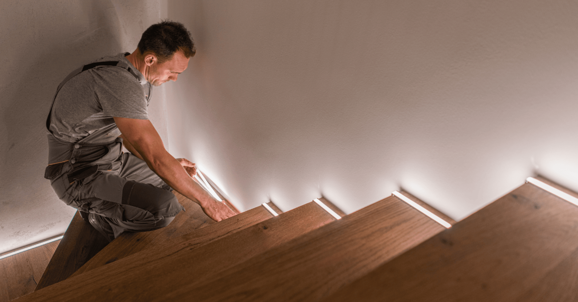 Ein Mann in Arbeitskleidung bringt LED-Lichtbänder am Rand einer Holztreppe an, wodurch die Stufen vor einer weißen Wand beleuchtet werden.