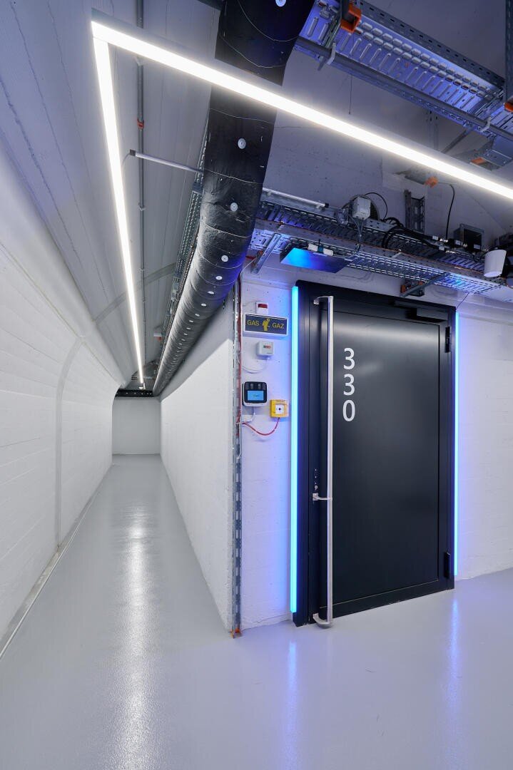 Ein moderner, schlichter Flur mit weißen Wänden und weißem Boden, freiliegenden Rohren und Kabeln an der Decke sowie einer großen schwarzen Tür mit der Aufschrift „330“, die an der Seite von blauen Lichtern beleuchtet wird.