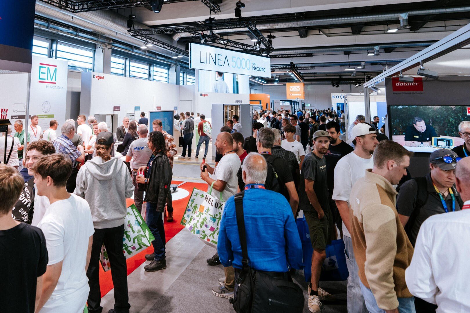 Eine große Menschenmenge bewegt sich durch eine Ausstellungshalle, in der sich im Hintergrund verschiedene Unternehmensstände, Banner und Ausstellungsstücke sehen lassen.
