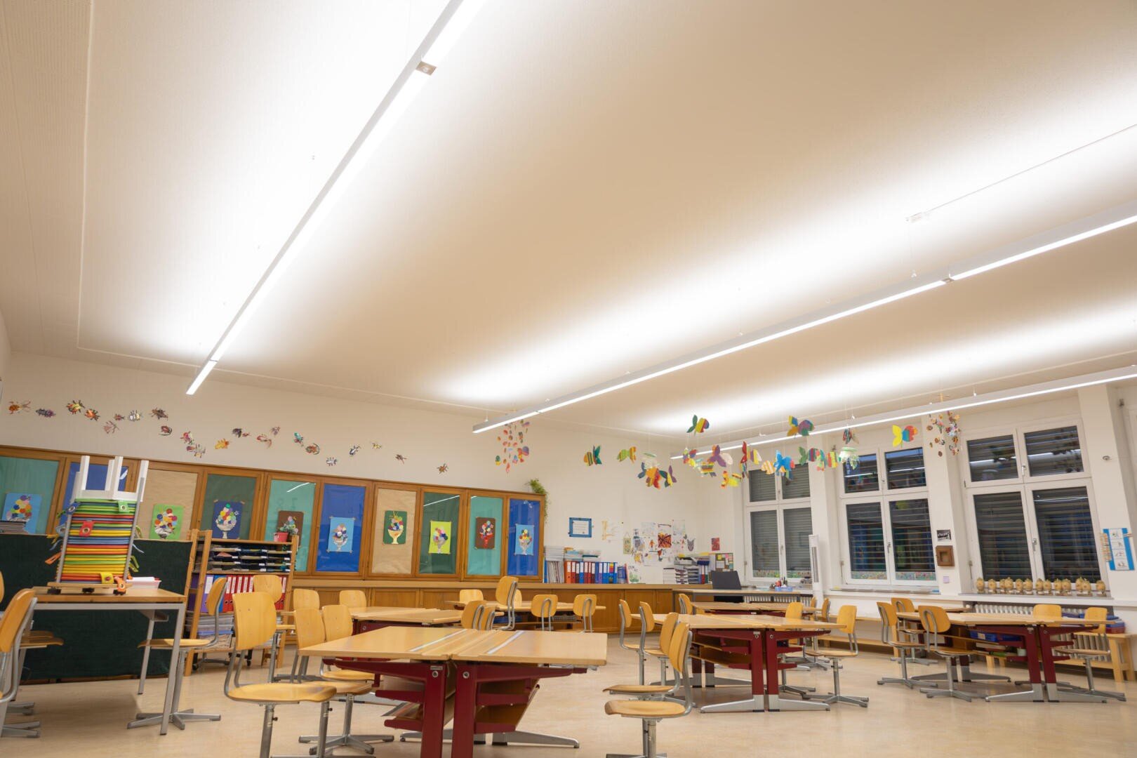 Une salle de classe vide et bien éclairée, avec des chaises et des tables en bois, des œuvres d'art colorées accrochées aux murs et des décorations en papier suspendues au plafond. De grandes fenêtres laissent entrer la lumière naturelle.