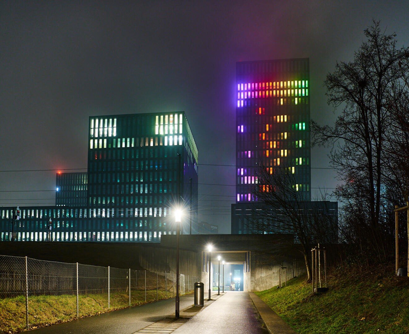 Eine Nachtszene zeigt einen Weg, der zu zwei modernen Hochhäusern führt. Die Gebäude sind mit bunten Lichtern in einem Rastermuster beleuchtet, während Straßenlaternen den Weg erhellen und rechts davon kahle Bäume stehen.