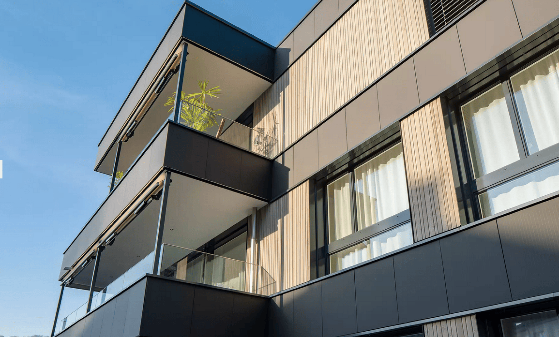 Modernes Mehrfamilienhaus mit großen Fenstern, Holzpaneelen und gläsernen Balkongeländern. Auf einem der oberen Balkone ist unter blauem Himmel eine Topfpflanze zu sehen.