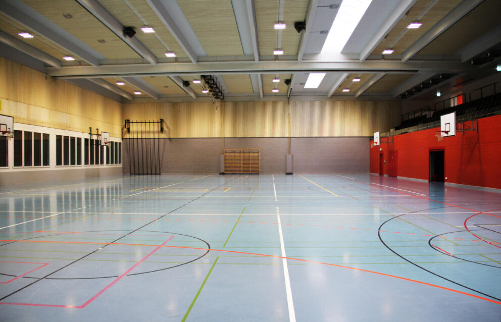 Große, leere Indoor-Turnhalle mit einem blauen, polierten Boden, der für verschiedene Sportarten markiert ist, Basketballkörben an beiden Enden, einer hohen Decke mit Lichtern und Sitzstufen entlang einer Seite.