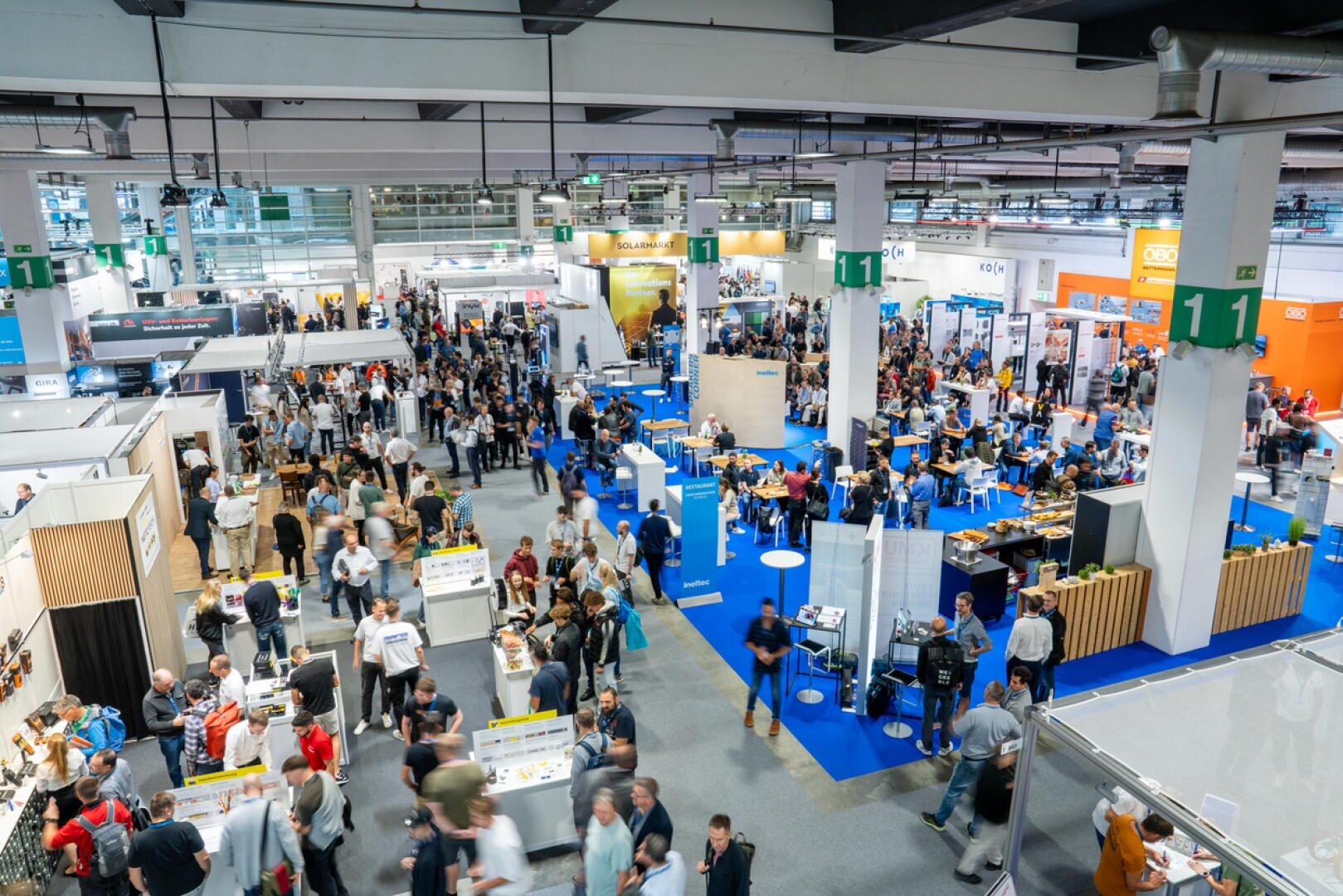 Eine große, belebte Kongresshalle, in der viele Menschen herumlaufen und verschiedene Stände besuchen. Blaue Teppiche säumen die Gänge, und die Aussteller stellen Produkte und Banner an ihren Ständen aus. Der Raum ist hell erleuchtet und lebendig.