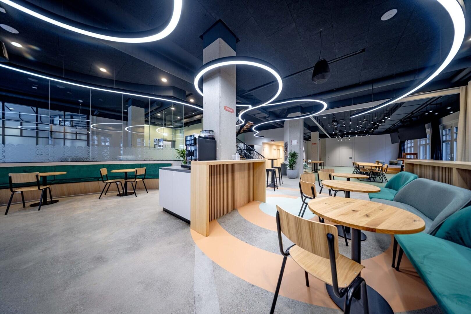 Modernes, geräumiges Café mit Holztischen, Stühlen, gepolsterten Sitzen in Petrol und einzigartigen runden LED-Deckenleuchten. Der Raum verfügt über große Fenster, eine Kaffeetheke und eine minimalistische Einrichtung.