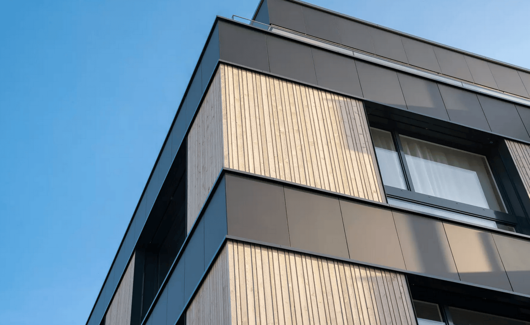 Eine moderne Gebäudefassade mit vertikalen Holzlatten und glatten schwarzen Metallpaneelen, mit großen Fenstern vor einem klaren blauen Himmel.