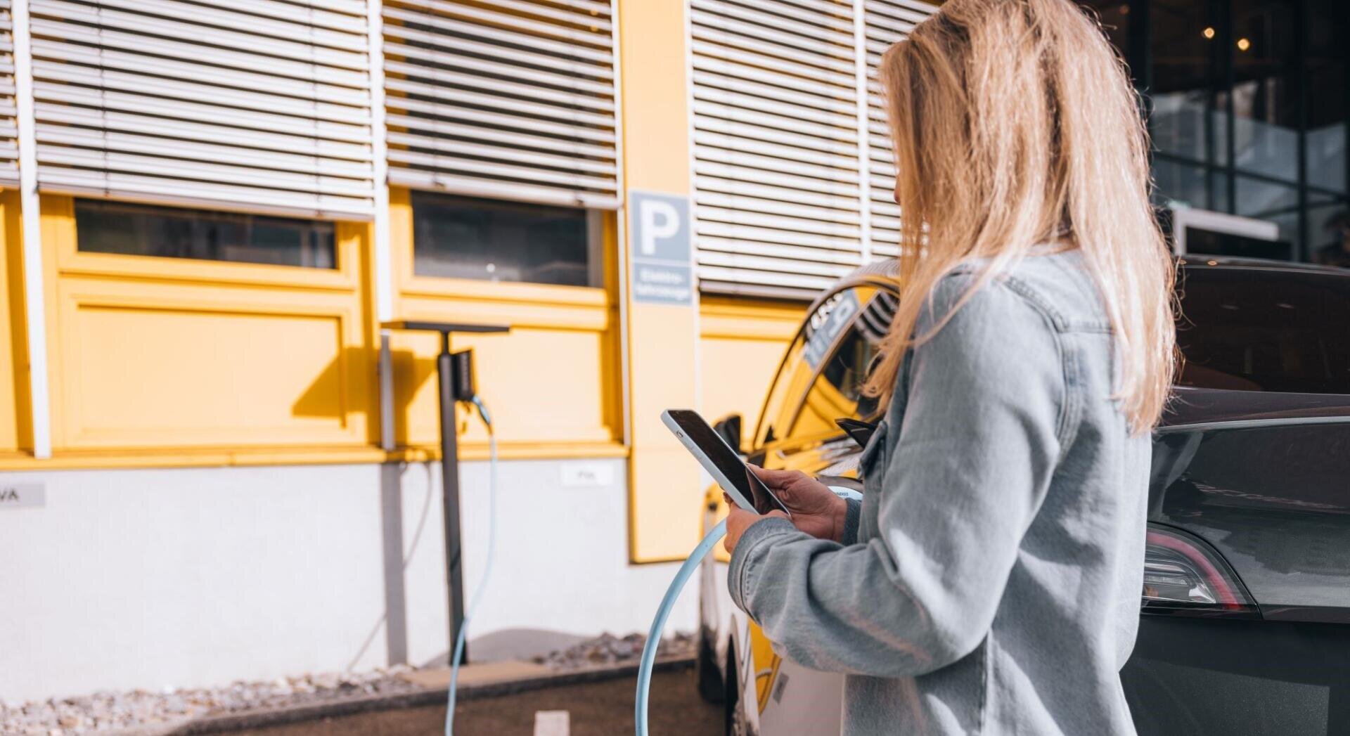 Une femme se tient à côté d'une voiture électrique près d'une borne de recharge, devant un bâtiment jaune ; elle tient un téléphone à la main et branche le câble de recharge sur la voiture.