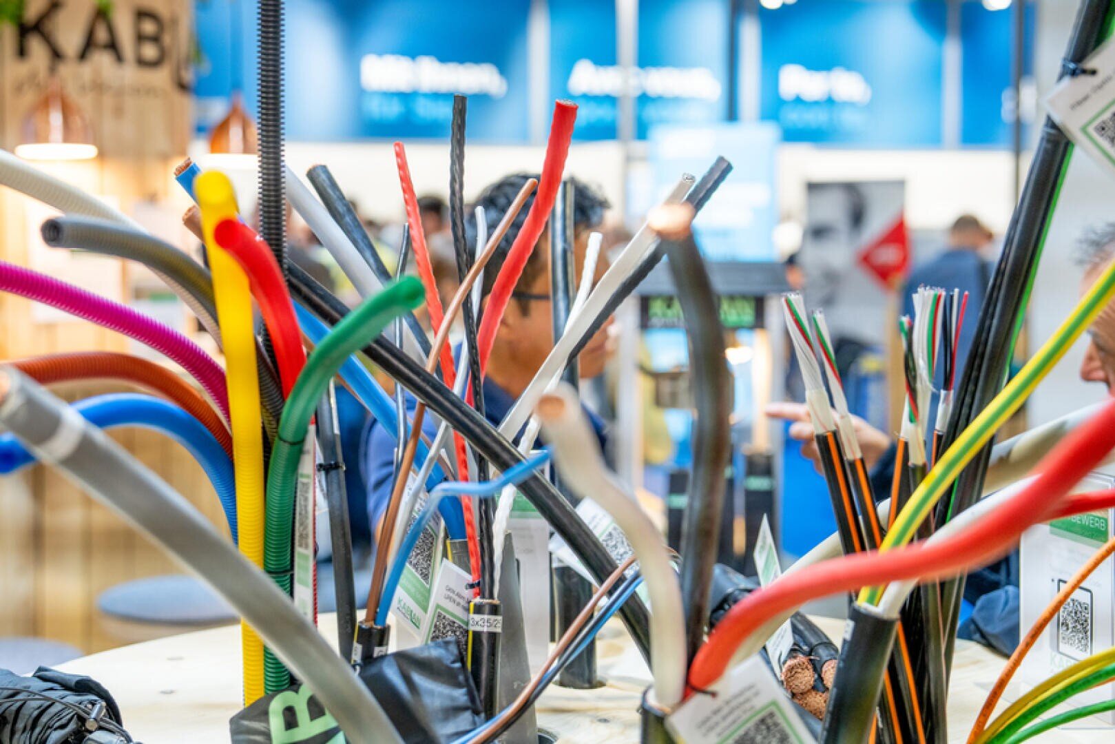 Ein Display mit verschiedenen bunten elektrischen Kabeln und Drähten, die aufrecht auf einem Tisch auf einer Messe stehen, mit verschwommenen Menschen und blauen Schildern im Hintergrund.