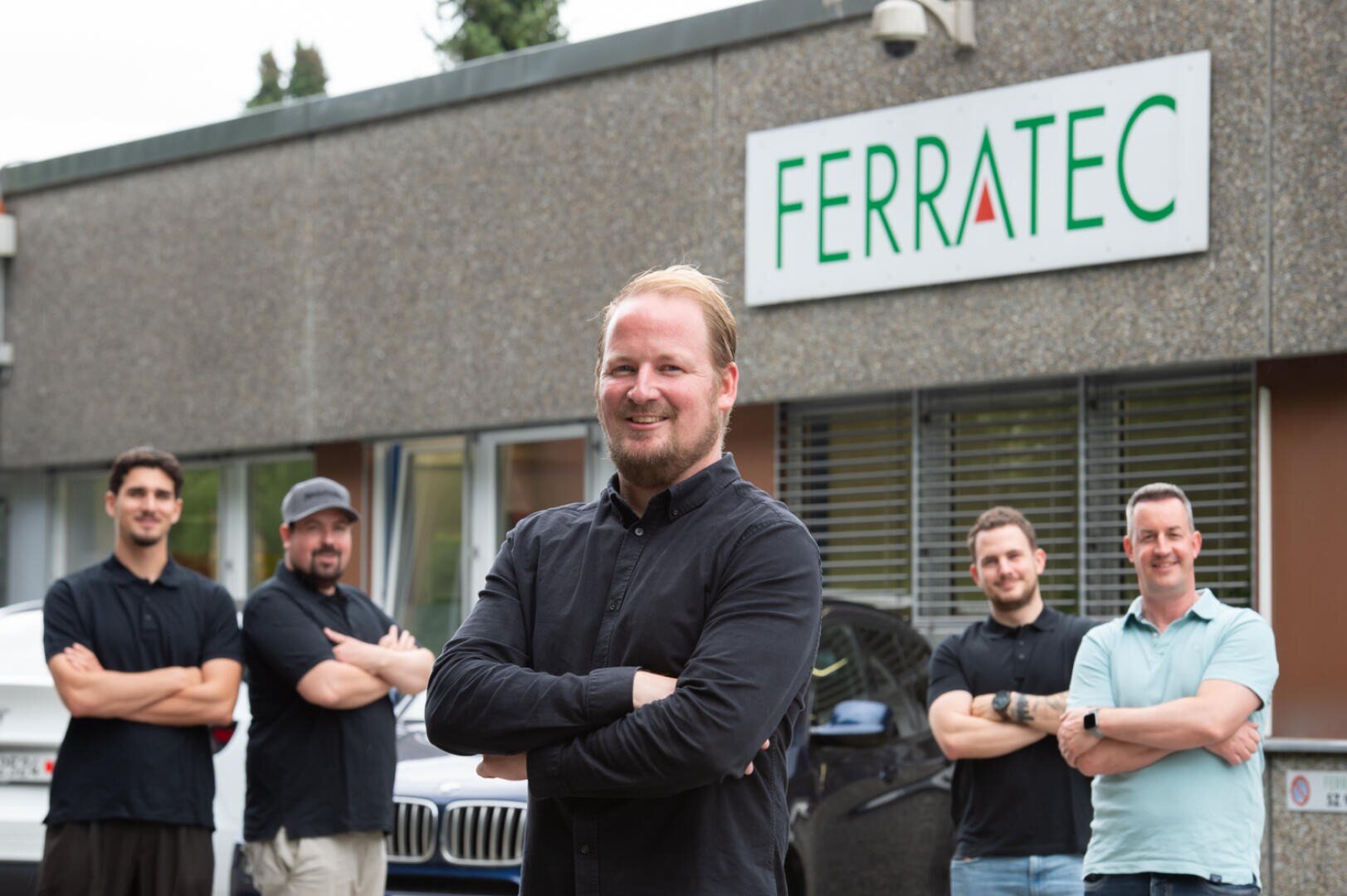 Fünf Männer stehen lächelnd und mit verschränkten Armen vor einem Gebäude mit einem FERRATEC-Schild. Der Mann ganz vorne steht in der Mitte, während die anderen vier hinter ihm stehen und leicht auseinander stehen.